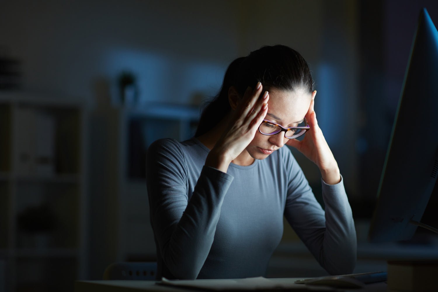 A woman sits at a desk, visibly stressed, holding her temples while looking at a computer screen in a dimly lit room.