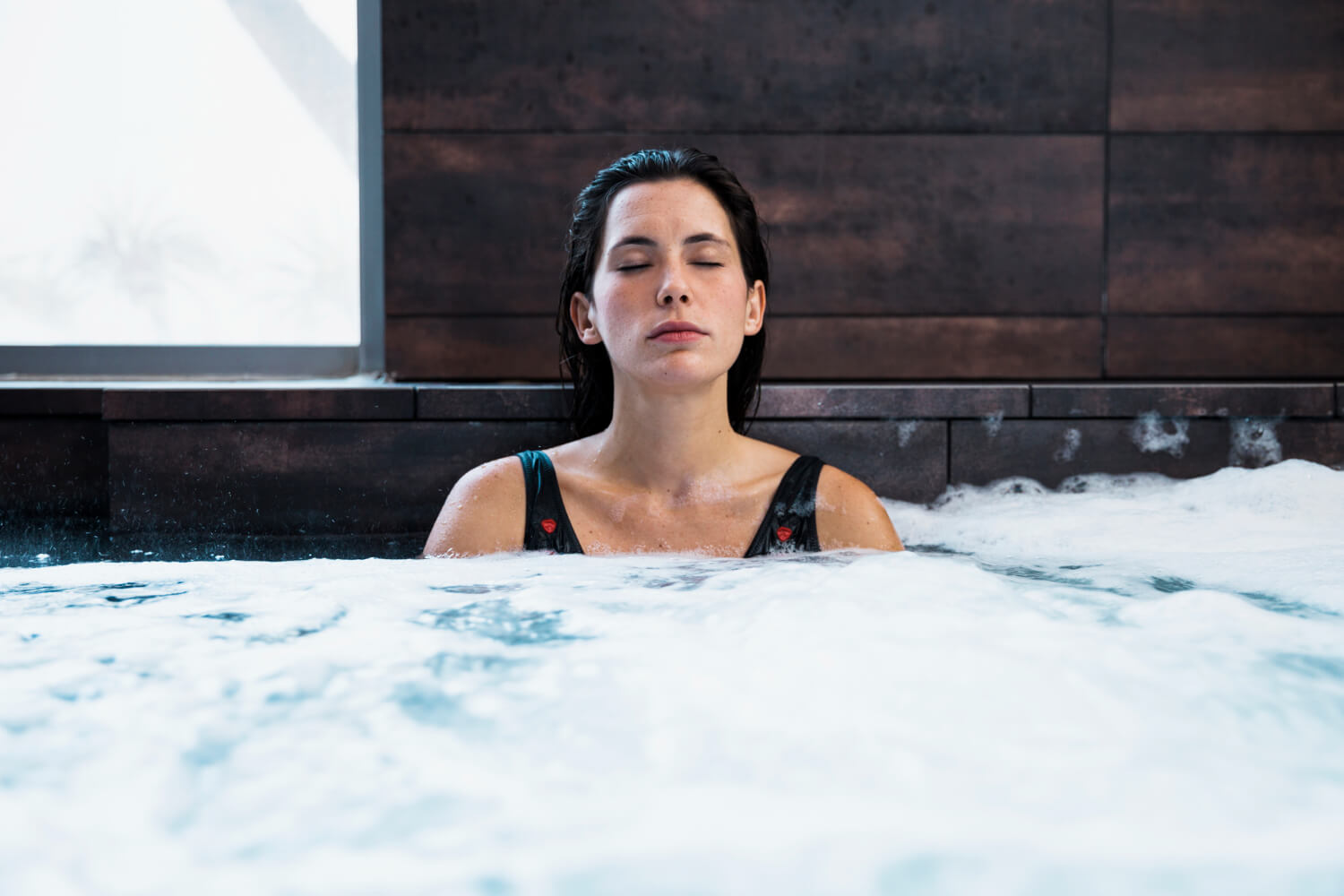 A woman in a swimsuit meditates in an ice tub with her eyes closed, surrounded by modern dark wood paneling.