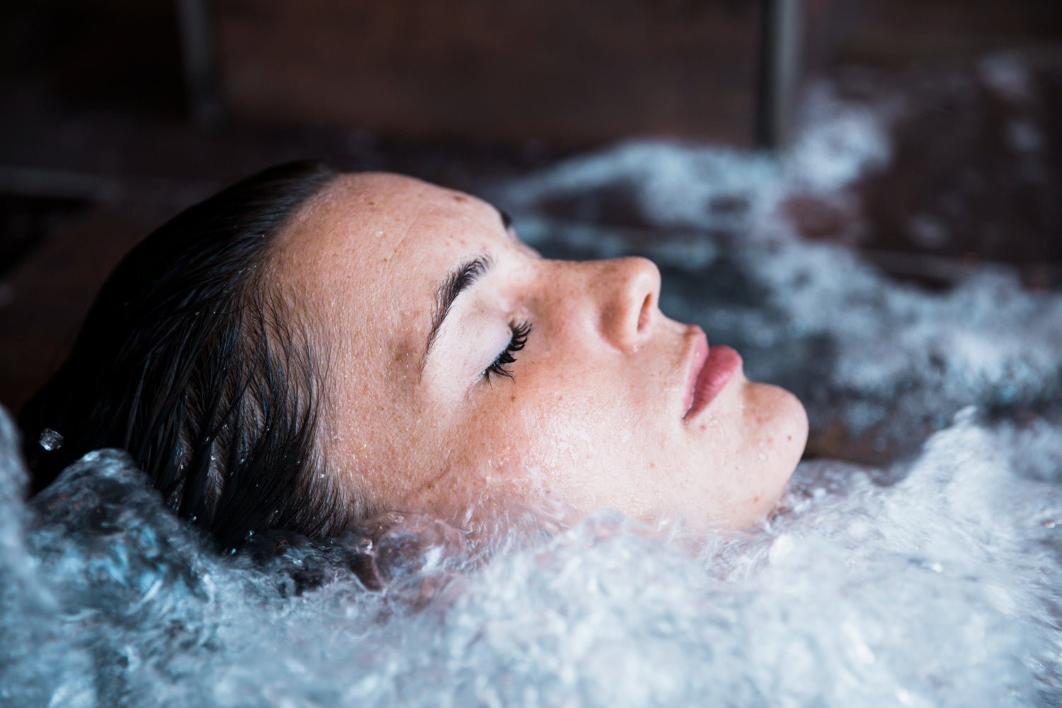 A woman relaxes in a bubbling indoor ice tub, her face serene and slightly submerged, with water ripples surrounding her.